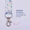 Das Bild zeigt einen Metallkarabiner, der an einem Stoffband mit Blumenmuster befestigt ist, sowie einen Text, der seine Verwendung zur Befestigung von Namensschildern oder Accessoires beschreibt.