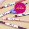 Das Bild zeigt mehrere bunte Stifte und Marker in Rosa- und Lilatönen, die jeweils mit 'INES L.' auf einer Holzoberfläche beschriftet sind.