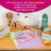 Das Bild zeigt eine schulszenische Szene mit bunten Buntstiften, die mit Namen beschriftet sind, einer Wasserflasche, einem Behälter mit Snacks und dekorativen Gegenständen wie Murmeln und Holzklötzen. Auf einem Tisch liegen Papiere in verschiedenen Farben ausgebreitet und an der Wand sind Zeichnungen zu sehen.