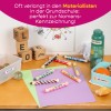 Das Bild zeigt einen Schulpult mit verschiedenfarbigen Buntstiften, die mit 'Léo Martin' beschriftet sind, Buchstabenblöcken, einem Behälter mit Murmeln, einer Wasserflasche und einer Snackbox mit einer Orange und einem Stück Brot. Im Hintergrund gibt es auch farbige Papierblätter und eine Zeichnung.