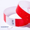 Das Bild zeigt ein rotes Armband mit einem weißen Abschnitt, der einige Markierungen aufweist, was darauf hinweist, dass es wasserdicht ist. Auf dem Armband befinden sich Wassertropfen.