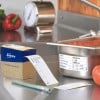 Das Bild zeigt eine Küchenszene mit einem Behälter Bolognese-Soße, einer Schachtel Avery-Etiketten, einem Stift und frischem Gemüse wie einer roten Paprika und einer Tomate.