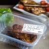 Ein Bild eines Plastikbehälters mit der Beschriftung "Kotelett in Senfkruste" datiert auf den 25.08.17, neben einem Teller mit gegrilltem Fleisch und Tomaten.