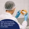 Eine Person in Schutzkleidung hält einen Behälter mit Lebensmitteln, der ein Etikett trägt. Die Umgebung scheint eine saubere, gereinigte Küchenumgebung zu sein, die die Einhaltung von Gesundheitsvorschriften betont.