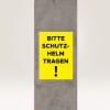 Das Bild zeigt ein gelbes Schild an einem Betonpfeiler mit der Aufschrift „BITTE SCHUTZ-HELM TRAGEN!“, was „Bitte einen Helm tragen!“ bedeutet und auf eine Sicherheitsanforderung hinweist.