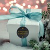 Das Bild zeigt eine weiße Geschenkbox, die mit einem blau-weiß gestreiften Band gebunden ist und auf einer schneebedeckten Oberfläche liegt. Sie hat einen runden Anhänger, auf dem 'Wir wünschen frohe Weihnachten' steht. Der Hintergrund ist weich verschwommen mit warmen, leuchtenden Lichtern.