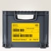 Das Bild zeigt eine graue Lagerkiste, die mit gelben Etiketten versehen ist. Die Etiketten enthalten Informationen wie 'Werk Bayern', einen Barcode und Produktdetails. Die Kiste scheint für Organisations- oder Versandzwecke verwendet zu werden.