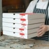 Eine Person in einer Kochuniform hält gestapelte Pizzakartons, die mit verschiedenen Namen und Bestellnummern beschriftet sind.