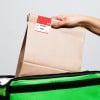 Eine Person hält eine braune Papiertüte mit einem Etikett darauf, das über einer grünen isolierten Liefer- bzw. Thermotasche platziert ist. Das Etikett enthält Details zur Bestellung.
