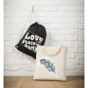 Das Bild zeigt zwei Taschen vor einer weißen Backsteinwand. Ein schwarzer Turnbeutel mit den Worten 'Love, Peace, Party' hängt dort. Daneben zeigt eine beige Tragetasche ein dekoratives Federdesign.