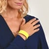 Das Bild zeigt eine Frau mit blondem Haar, die ein dunkelblaues Hemd trägt. Sie ist mit einer goldenen Halskette geschmückt und trägt ein leuchtend gelbes Armband am Handgelenk, wobei ihre Hand sanft auf ihrer Schulter ruht.
