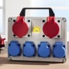 Das Bild zeigt eine Stromverteilungsbox mit zwei roten runden Steckdosen oben und vier blauen runden Steckdosen unten. Sie ist mit einem Griff für den Transport ausgestattet und besitzt einen gelben Zertifizierungsaufkleber, der anzeigt, dass sie den Sicherheitsstandards entspricht.