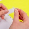 Das Bild zeigt die Hände einer Person, die einen Papier- oder Aufkleberstreifen von einem Blatt abziehen, mit einem leuchtend gelben Hintergrund und gedruckten Designs auf dem Streifen.