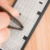 Das Bild zeigt eine Hand, die mit einer Pinzette Buchstaben aus einem Satz kleiner weißer Plastikplättchen entnimmt, die in Reihen angeordnet sind, mit einer hölzernen Oberfläche darunter.