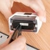 Das Bild zeigt eine Person, die mit einer Pinzette ein kleines Objekt in einem Handgerät oder Stempel handhabt. Im Hintergrund ist eine Holzoberfläche zu sehen, auf der weitere Stempel oder Buchstaben sichtbar sind.