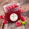 Das Bild zeigt ein Glas Himbeermarmelade mit frischen Himbeeren und Minzblättern, die darum herum platziert sind und auf einer Holzoberfläche liegen. Das Glas hat ein Etikett mit der Aufschrift 'Himbeermarmelade' und weist darauf hin, dass es hausgemacht ist.
