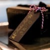 Das Bild zeigt ein hölzernes Geschenkanhänger mit dem goldenen Text 'Frohes Fest'. Der Anhänger ist mit festlichen Symbolen wie einer Schneeflocke, Glocken, einer Kerze und immergrünen Zweigen verziert und mit rotem und weißem Garn gebunden.