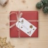 Das Bild zeigt eine wunderschön verpackte rote Geschenkschachtel mit einem transparenten Band und einem Geschenkanhänger, der einen handgeschriebenen Namen enthält. Auf dem Anhänger befinden sich dekorative Elemente wie Sterne. In der Nähe gibt es eine kleine immergrüne Dekoration und einen sternförmigen Anhänger, die alle auf einer hölzernen Oberfläche platziert sind.