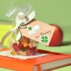 Das Bild zeigt eine kleine Geschenktüte, gefüllt mit bunten Gummibärchen, gebunden mit einem Band und einem Anhänger mit der Aufschrift 'Viel Glück zur Prüfung'. Sie liegt auf einem orangefarbenen Buch, mit einem Apfel im Hintergrund vor einem grünen Hintergrund.