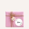 Das Bild zeigt ein wunderschön verpacktes rosa Geschenkbox, verziert mit Schnur und einer kleinen Blume. Ein runder Anhänger mit dem Namen 'Anna' ist an das Paket angebracht.