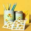 Das Bild zeigt zwei verzierte Blechdosen, die Buntstifte halten. Die Dosen sind mit Sonnenblumenaufklebern und grünen karierten Bändern verziert und vor einem leuchtend gelben Hintergrund platziert. Neben den Dosen liegen Bögen mit Sonnenblumenaufklebern, die der Szene eine fröhliche und künstlerische Atmosphäre verleihen.