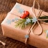 Das Bild zeigt ein wunderschön verpacktes Geschenk in braunem Papier, verziert mit einem natürlichen Kordelbogen, grünen Blättern, roten Beeren und blauen sternförmigen Ausschnitten.