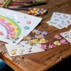 Das Bild zeigt einen kreativen Arbeitsbereich mit bunten Zeichnungen und verschiedenen Aufklebern. Ein Regenbogen ist auf Papier gemalt und mit glänzenden Edelsteinen dekoriert. Es gibt mehrere Bögen mit Aufklebern von Cartoon-Charakteren, Herzen und Blumen, zusammen mit Buntstiften und verstreuten Süßigkeiten auf einem Holztisch.
