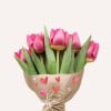 Ein Strauß rosa Tulpen in Papier mit dekorativen Herzen gewickelt, vor einem hellen Hintergrund platziert.