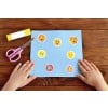 Das Bild zeigt ein blaues Papier mit bunten Sonnenstickern darauf, eine rosafarbene Schere und einen Klebestift, die auf einem Holztisch liegen. Die Hände eines Kindes sind sichtbar, eine Hand berührt das Papier mit den Stickern, während die andere die Schere hält.